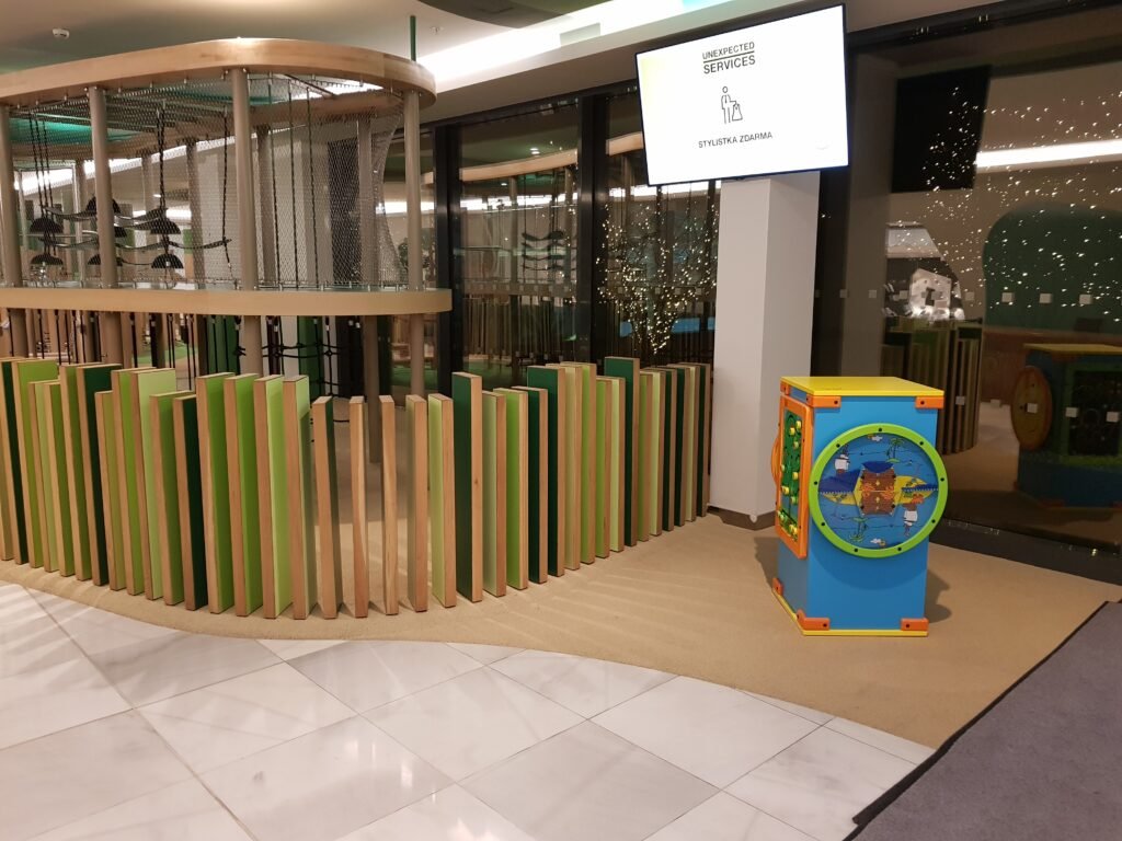 Vnitřní dětský koutek s kruhovou dřevěnou a zelenou bariérou, lezeckou konstrukcí, interaktivním hracím panelem a pískovou podlahou. V pozadí je vidět velké okno a digitální nápis.