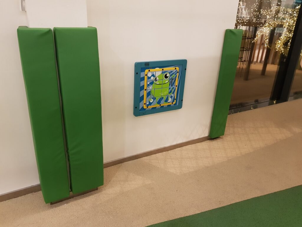 Stěna se dvěma svislými zelenými polstrovanými chrániči a čtvercovou interaktivní dětskou nástěnnou hračkou s bludištěm. Podlaha je béžová a zelená a vpravo je vidět skleněná stěna se světly.
