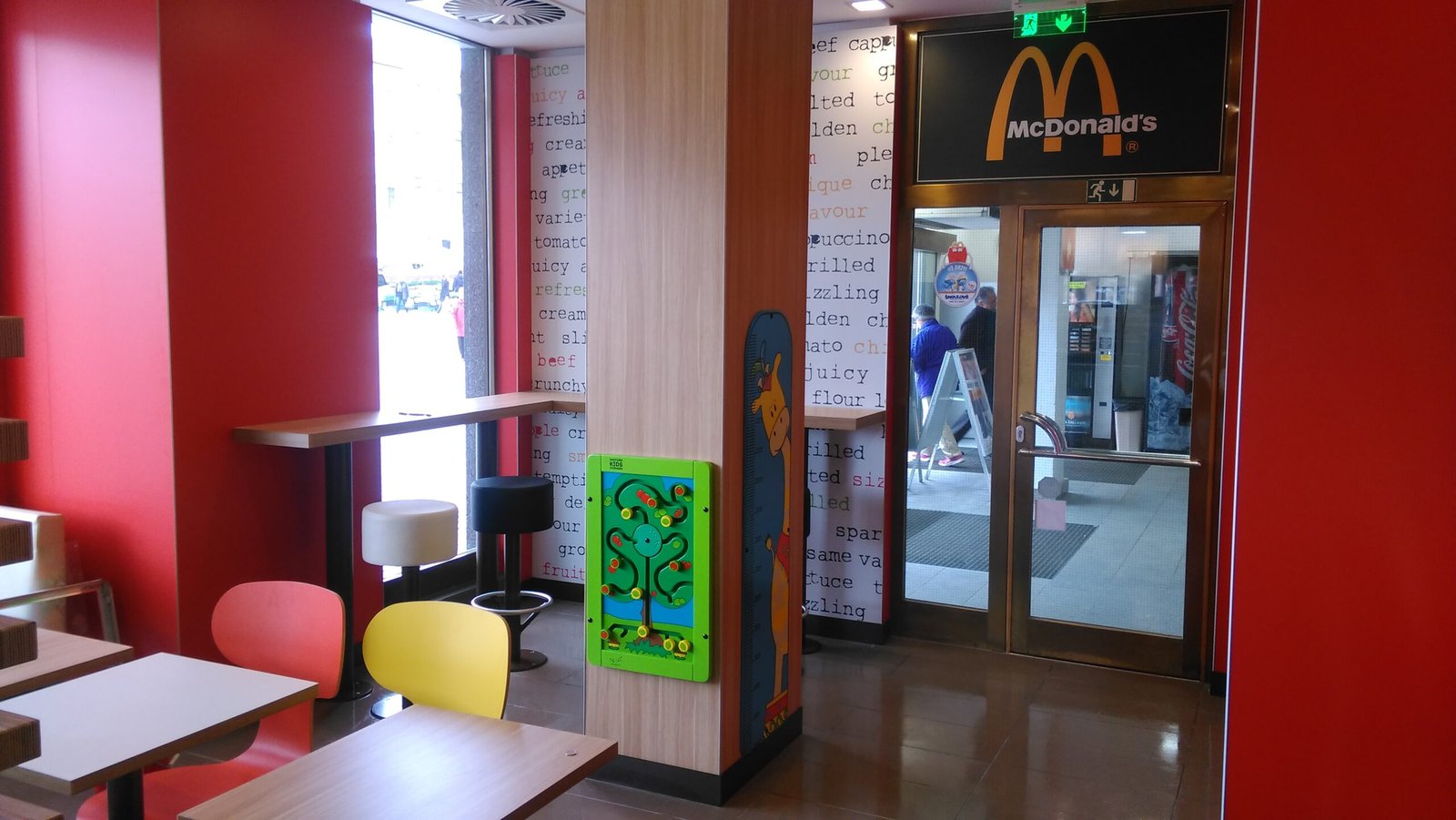 Interiér restaurace McDonald's s barevnými židlemi, malými stolky, dětskou nástěnnou hrou a skleněnými dveřmi s logem McDonald's nad nimi. Červené a dřevem tónované stěny vytvářejí světlý a příjemný prostor.