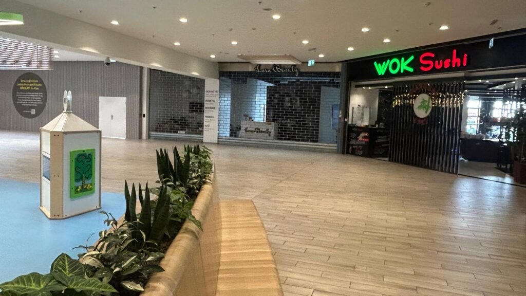 Převážně prázdná chodba obchodního centra se zavřeným obchodem uprostřed, lavičkou s rostlinami vlevo a osvětlenou restaurací WOK Sushi vpravo. Na podlaze je dlažba a prostor působí klidně.