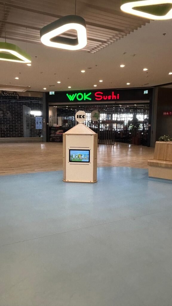V prázdném nákupním centru stojí robotický kiosek s dotykovou obrazovkou; v pozadí je zeleným a červeným neonem vyveden nápis restaurace WOK Sushi.