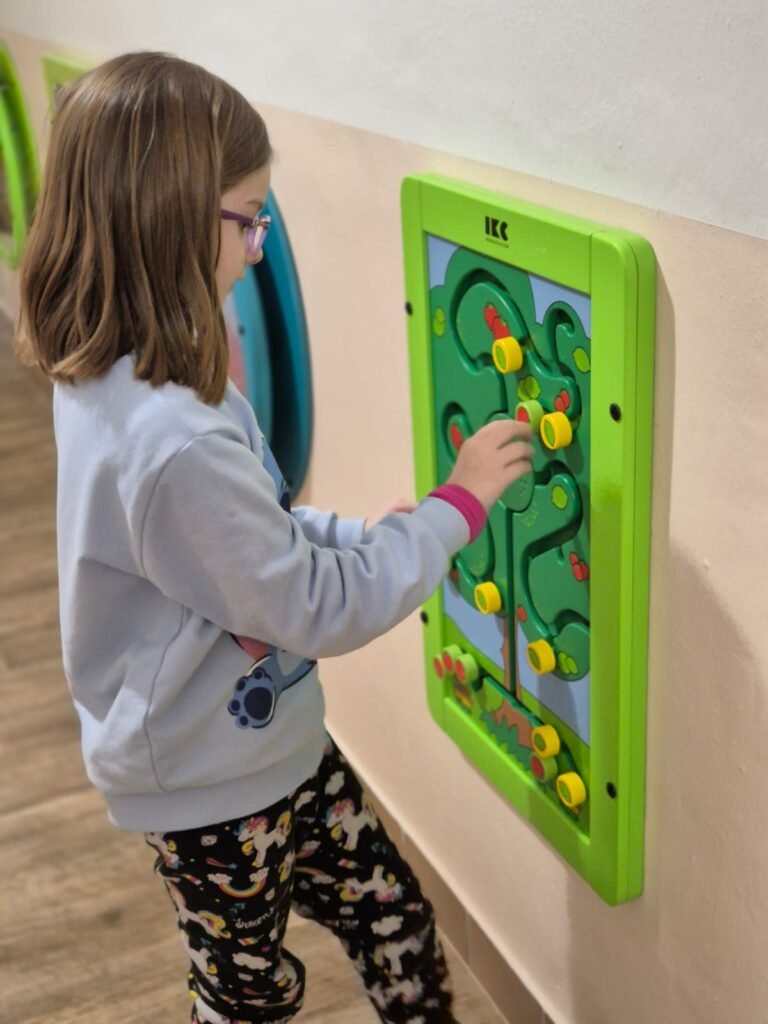 Mladá dívka s brýlemi si hraje s barevným nástěnným bludištěm a posouvá žluté a červené dílky po klikatých cestičkách. Na sobě má světle šedý svetr a vzorované kalhoty.
