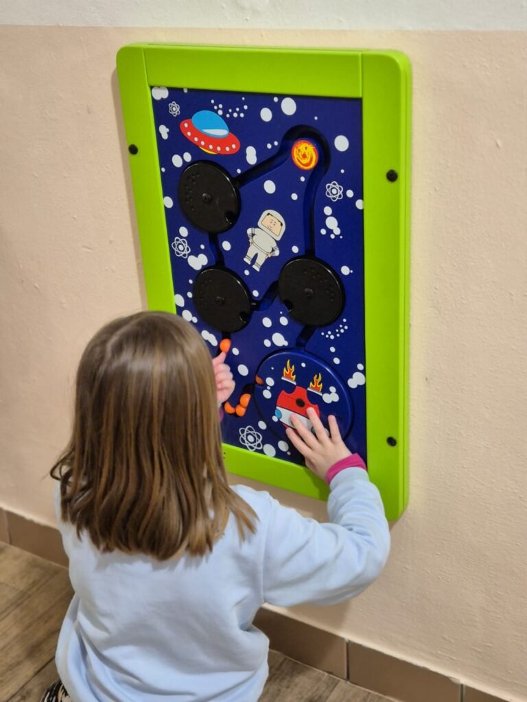 Dítě se světle hnědými vlasy a světle modrým svetrem si hraje s barevnou nástěnnou tabulí s vesmírnou tematikou, která obsahuje astronauta, UFO, planety a pohyblivé knoflíky.