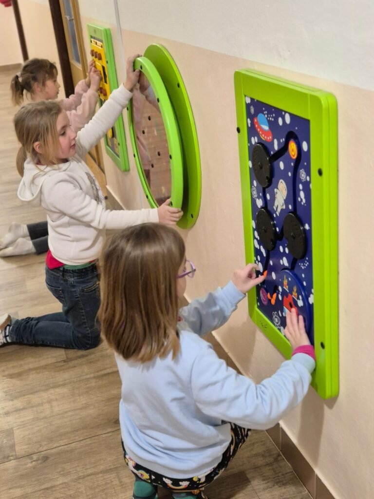 Tři malé děti klečí na dřevěné podlaze a věnují se barevným interaktivním panelům na stěně v chodbě. Panely obsahují hry a aktivity a děti vypadají soustředěně a zaujatě.